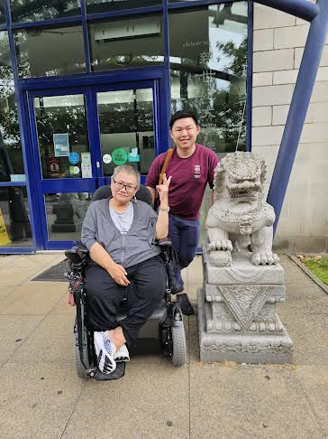 two East Asians, one  standing and the other in a wheelchair. they are next to a stone lion