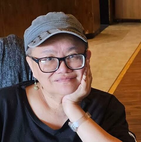 East Asian with blue cap and dark rimmed glasses. She/they is/are wearing a black tee shirt, her head is resting on left arm.
