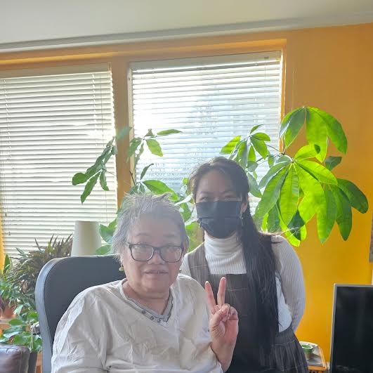 2 East Asian women - one with white tee shirt, seated with 2 fingers up, the one at the back. standing, masked with long hair, white polo neck wth grey pinafe. theres a big plant behind them, window with blinds and a bright yellow wall.